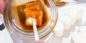 A person is dipping a marshmallow into a jar of caramel sauce using a stick. Several white marshmallows are visible on a plate in the background. A spoon covered in caramel sauce lies next to the plate.
