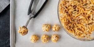 An image showing a tray with six cheese and meat mixture balls arranged on a baking sheet. A bowl with more of the mixture is next to the tray, and a scoop ladle with a portion of the mixture is positioned between the bowl and the balls on the tray.