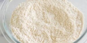A clear glass bowl filled with white, finely ground flour. The texture appears soft and powdery, with subtle indentations and a light, uneven surface. The bowl is placed on a white countertop, highlighting the flour's pale color.