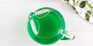 A top-down view of a glass pitcher filled with green liquid, placed on a white surface. To the right of the pitcher, partially visible, is a white cloth with green leaf patterns.