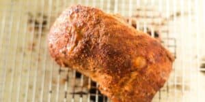A cooked, seasoned pork roast sits on a metal rack. The roast has a crispy, golden-brown crust, suggesting it was roasted or grilled to perfection. The metal rack is placed on what appears to be a baking sheet.