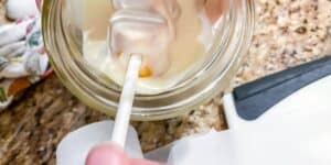 A hand dips an orange and white food item on a stick into a clear jar filled with a creamy, white substance. The jar is on a granite countertop, with a cutting board and patterned cloth partially visible in the background.