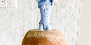A decorative figurine of Elsa from Disney's Frozen is placed upside down in a large loaf of bread. The background features a white herringbone-patterned wall with a visible power outlet on the left.