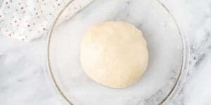 A ball of dough rests in a clear glass bowl on a white marble surface. A white cloth with small colorful dots is partially visible in the top left corner of the image.
