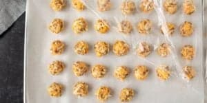 A baking sheet lined with parchment paper holds multiple rows of unbaked cheese balls. A piece of plastic wrap partially covers the cheese balls, indicating they are likely being prepared for baking or freezing. A black surface is visible around the baking sheet.