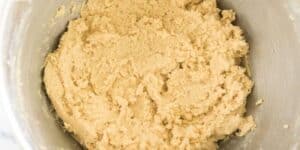 A close-up of cookie dough in a metallic mixing bowl. The dough is a light beige color and has a rough, slightly crumbly texture.