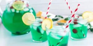 Three glasses of vibrant blue-green punch with ice cubes, garnished with lemon slices and cherries, and striped red and white straws. There's a matching pitcher filled with the same punch in the background. The scene appears bright and fresh.