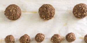 A baking sheet lined with parchment paper holds various sizes of raw, brown falafel balls, evenly spaced. The larger falafel are in the center row, while the smaller ones are lined up in the bottom row, ready to be baked.