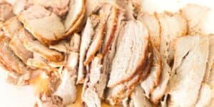 Sliced, cooked pork lies arranged on a white surface. The meat appears tender with a light brown crust on the edges, hinting at its roasted preparation. Juices are visible around some slices, indicating its succulent texture.