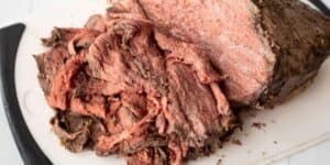 A sliced roast beef is shown on a white cutting board. One end of the beef is intact, while the other end has several slices that reveal the pink, tender interior. The darker, crusted exterior showcases the seasoning and cooking technique used.
