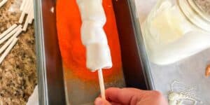 A hand is holding a cake pop coated in white chocolate, while dipping it into a shallow tray of orange sprinkles. On the right, there is a jar filled with white chocolate, and on the left are several white cake pop sticks.
