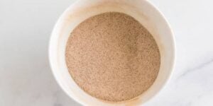 A white bowl filled with a fine, light brown granulated substance, which appears to be yeast or a similar powder, is placed on a smooth white surface.