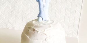 A white frosted cake topped with a small, light blue figure wearing a suit. The background features a subtle chevron pattern. The overall scene is minimalist and the cake appears to be the main focus.