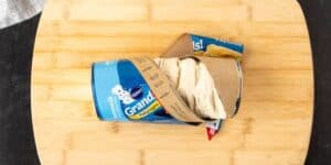 A tube of refrigerated Pillsbury Grands! Buttermilk biscuit dough is partially opened on a wooden cutting board. The dough is starting to spill out from the tube’s packaging.