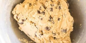 Close-up of raw cookie dough with chocolate chips in a silver mixing bowl. The dough has a light beige color with visible chunks of dark chocolate, ready for baking. The texture appears soft and slightly crumbly.