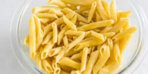 A clear glass bowl filled with cooked penne pasta, with each piece of pasta showcasing its characteristic cylindrical shape and diagonal cuts at the ends. The pasta appears to be plain with a light yellow hue, indicating it's simply boiled without sauce.