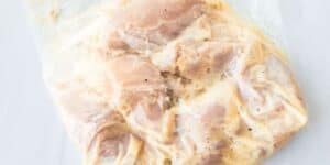 A clear plastic bag containing pieces of marinated raw chicken. The marinade appears to be a creamy, opaque liquid, possibly with seasonings. The bag is sealed and placed on a white surface.