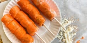 Four corn dogs coated with a bright orange breading sit on a white paper plate. To the right of the plate, there's a mess of melted cheese and orange crumbs on a piece of parchment paper. The corn dogs appear crispy and crunchy.