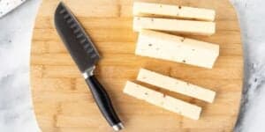 A knife with a black handle rests on a wooden cutting board next to several rectangular slices of cheese. The cheese has small specks, possibly herbs or spices, embedded in it. The background is a marble countertop.