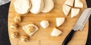 A wooden cutting board with pieces of dough at various stages of preparation, including flat rounds, quartered sections, and small balls of dough. Some of the balls are coated with a mixture of shredded cheese and nuts. A knife lies to the right.
