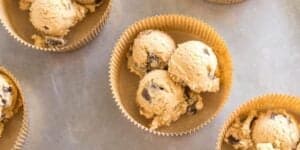 Scoops of chocolate chip cookie dough are placed in brown paper baking cups on a metallic surface. Additional baking cups with cookie dough are slightly visible in the background.