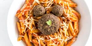 A plate of penne pasta in a rich tomato sauce, garnished with shredded cheese. Three breaded and seasoned meatballs are arranged on top, resembling Mickey Mouse's face. A small green leaf sits on the central meatball.