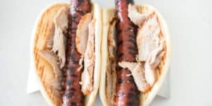 Two grilled sausages with char marks are placed inside flatbread wraps, each topped with slices of roast meat. The wraps are positioned side by side on a white surface, and the meat looks well-cooked with a slightly browned texture.