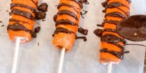 Three vibrant orange deep-fried corn dogs on sticks sit on parchment paper, drizzled with a generous amount of melted chocolate. A spoon hovers over one corn dog, adding more chocolate drizzle.