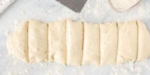 A piece of dough rolled into a flattened rectangle lies on a flour-dusted surface. The dough has been cut into evenly spaced vertical sections, ready for further preparation or baking. A metal scraper and a bowl with flour are visible in the background.