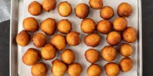 A metal tray is lined with paper towels and filled with multiple evenly spaced, golden-brown, round fried dough balls. The dough balls have a uniform color and texture, indicating they were freshly fried.