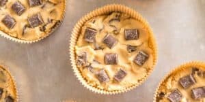 A close-up image of muffin liners filled with unbaked cookie dough. The dough is embedded with chocolate chunks. The liners are placed on a metallic baking tray, ready to be baked.
