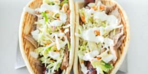 Two soft shell tacos filled with shredded pork, a vibrant mix of cabbage slaw, thinly sliced carrots, and drizzled with a creamy white sauce. The tacos are placed side by side on a metal taco holder against a light background.