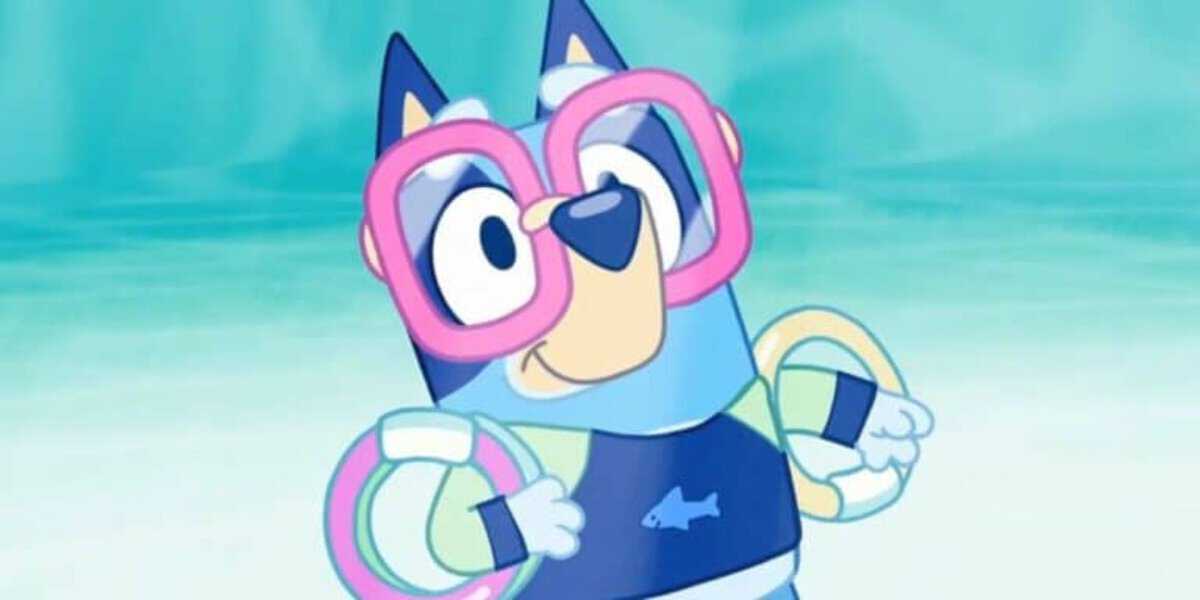 A cartoon Bluey blue heeler dog wears pink swim goggles, floaties on both arms, and a dark blue swimsuit with a fish logo, standing against a light blue background.