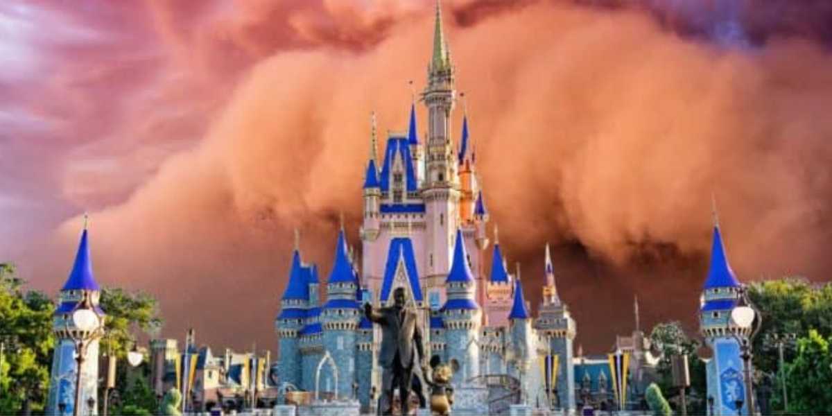 A towering, ornate castle with blue and gold spires stands prominently under a dramatic, vivid orange sky. In front of the castle, a statue of Walt Disney holding hands with Mickey Mouse is visible. Lush greenery and decorative lampposts surround the scene.