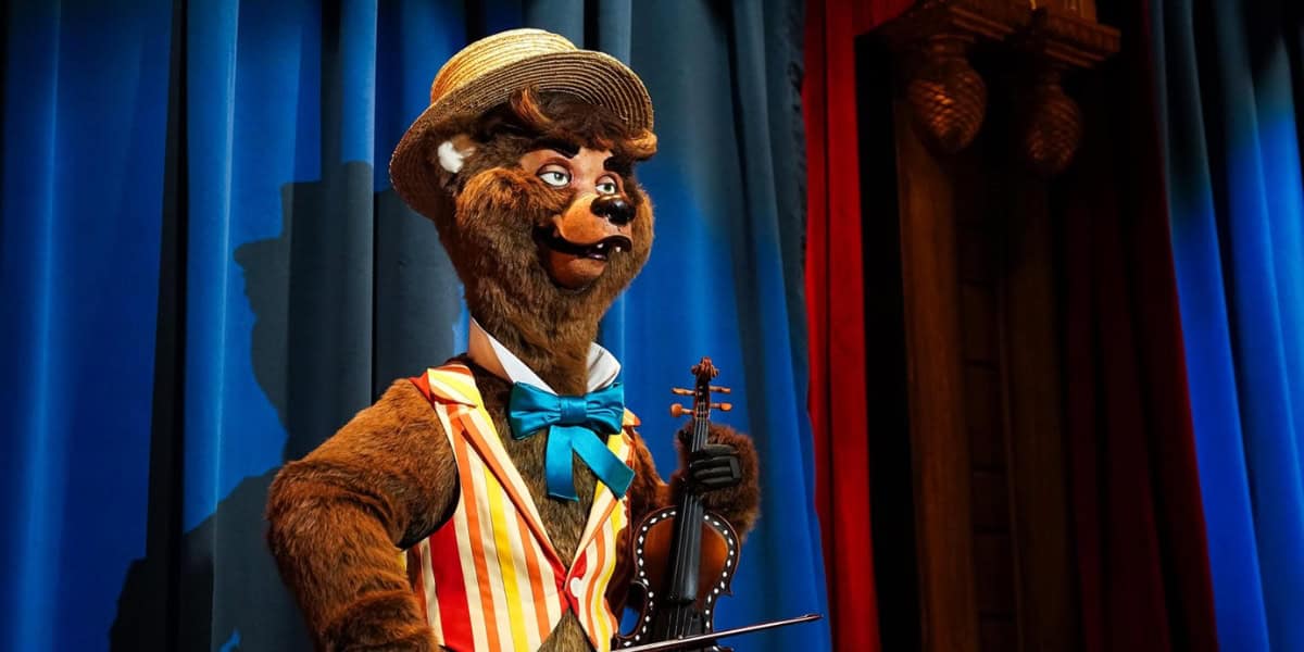A bear dressed in a colorful striped vest, blue bow tie, and straw hat stands against a backdrop of blue and red curtains, holding a violin and looking to the side. The bear has a cheerful expression and is part of an animated performance.