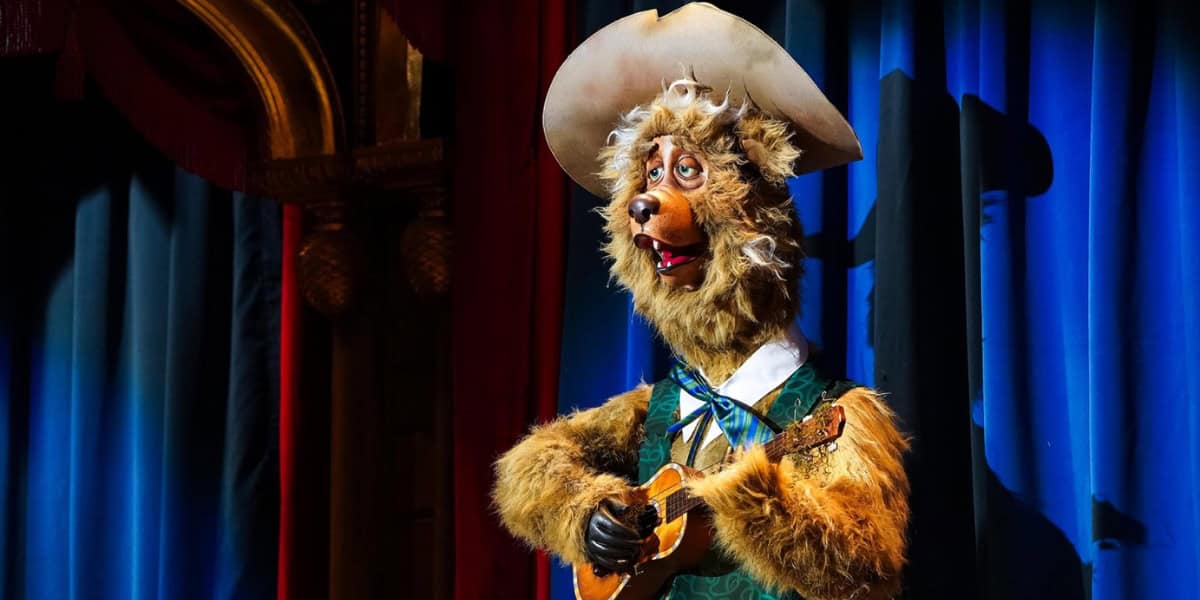 An animatronic bear dressed as a cowboy, wearing a wide-brimmed hat, green vest, and bow tie, plays a small guitar. The bear stands on a stage with blue curtains.