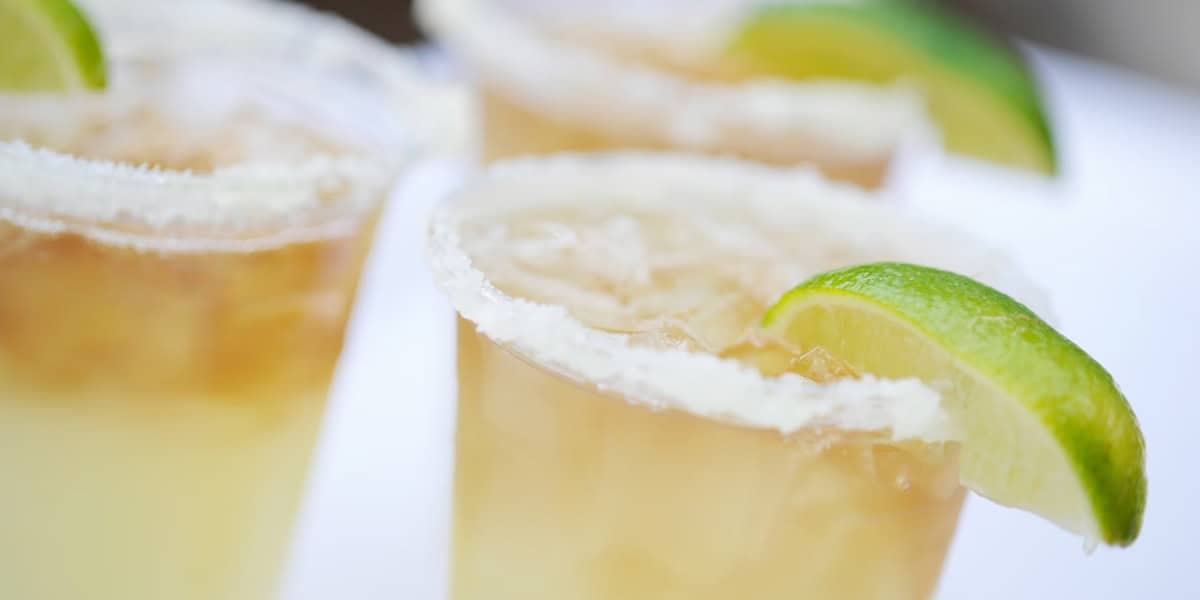 Close-up of three margaritas in clear plastic cups, each garnished with a salted rim and a vibrant lime wedge. The drinks are filled with ice, and the background is softly blurred, drawing focus to the refreshing beverages.