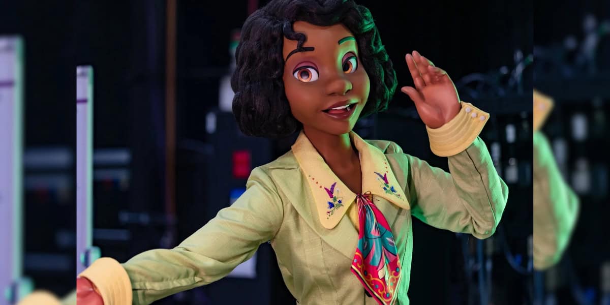 A life-like figurine of an animated character with a brown complexion, curly dark hair, and large expressive eyes is seen in the image. The character is wearing a light green blazer with a colorful scarf and flower designs on the collar, set against a blurry background.