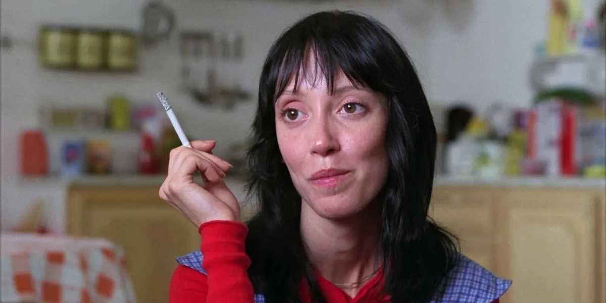 A person with long black hair is seen holding a cigarette in their left hand. They are wearing a red top with a blue checkered apron. The background shows a kitchen with various items on shelves and countertops. The expression on the person's face appears contemplative.