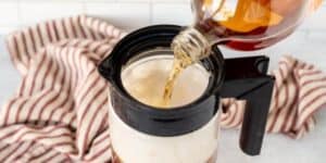 A hand pours maple syrup from a glass bottle into a jar of frothy milk. The jar sits on a countertop with a red and white striped cloth in the background.