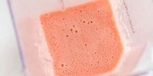 A blender jar filled with smooth, light pink liquid, likely a smoothie, is shown from above. The jar has a pink, frothy mixture with small bubbles on the surface, against the transparent walls of the blender.