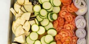 A baking tray is filled with sliced vegetables arranged in rows. From left to right, there are slices of eggplant, zucchini, tomatoes, and red onion. The tray is placed on a light-colored surface.