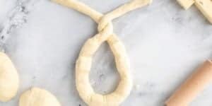 A partially twisted piece of dough in the process of being shaped into a pretzel on a marble countertop. Surrounding it are other dough pieces and a small rolling pin—it's the beginning stage of our Jalapeño Cheese Stuffed Pretzel recipe inspired by Disney.