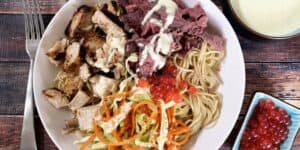 A Satu'li Bowls Recipe inspired by Pandora - World of Avatar features a bowl filled with grilled chicken pieces, shredded cooked meat with white sauce, cooked noodles, and a mix of shredded vegetables like carrots and bell peppers. Red fish roe is sprinkled on top, accompanied by a fork and additional sides around the bowl.