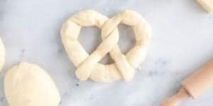 A raw pretzel-shaped dough placed on a marble surface, surrounded by additional dough pieces. A small wooden rolling pin is visible to the right. The scene is bright and minimal, evoking a cozy kitchen ready for creating a Jalapeño Cheese Stuffed Pretzel Recipe inspired by Disney magic.