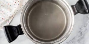 A stainless steel pot with black handles is filled with simmering water, emitting steam, as you prepare for a Jalapeño Cheese Stuffed Pretzel Recipe inspired by Disney. The pot sits on a gray countertop next to a white cloth with small, scattered red and blue dots.