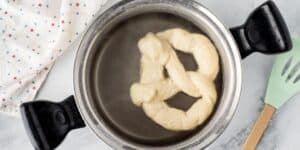 A twisted dough pretzel is boiling in a pot of water. The pot has black handles, and a mint green spatula is placed to the right side. A white kitchen towel with small colored patterns is partially visible to the left. This scene could be straight out of a Jalapeño Cheese Stuffed Pretzel recipe inspired by Disney.