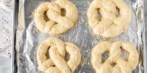 Four uncooked pretzels with shredded cheese sprinkled on top are placed on a baking sheet lined with aluminum foil. The baking sheet is slightly tilted, displaying the dough in preparation for baking. Inspired by a Disney favorite, these jalapeño cheese stuffed pretzels promise a delicious twist.