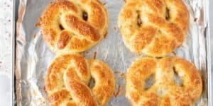 Four golden-brown pretzels, inspired by Disney's jalapeño cheese stuffed pretzel recipe, are topped with melted cheese and arranged on a foil-lined baking sheet. The freshly baked pretzels have a soft and warm appearance, with small cheese bits scattered around them on the foil.