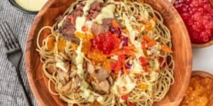 A wooden bowl filled with spaghetti, topped with slices of meat, mixed vegetables, and a variety of colorful popping boba pearls. A small cup of sauce is visible to the side, along with a gray napkin, fork, and spoon. Satu'li Bowls Recipe Inspired by Pandora - World of Avatar bring an otherworldly touch to this dish.